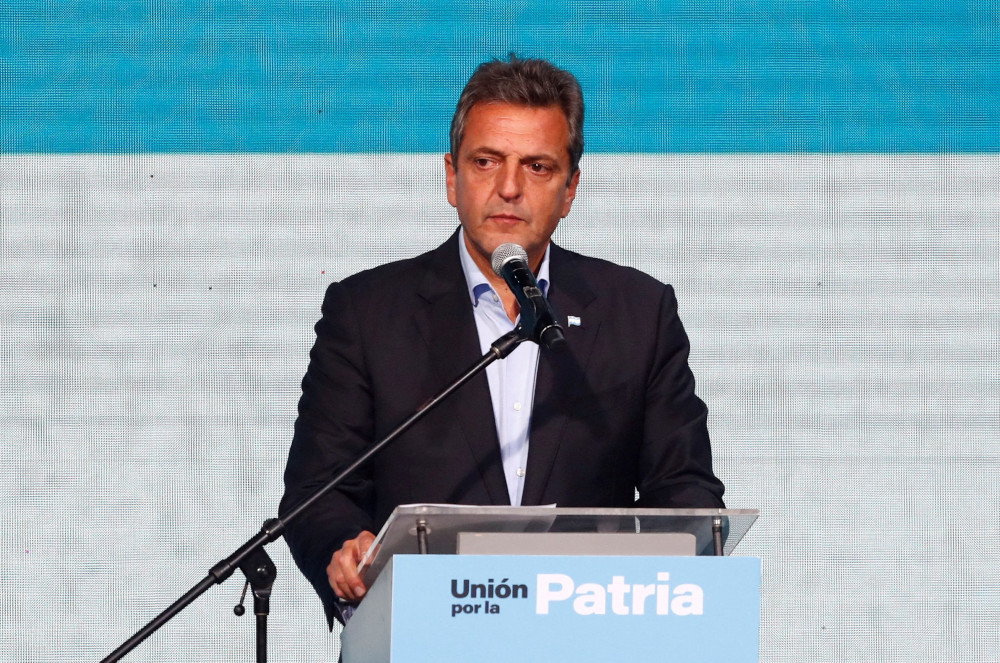 A light-skinned man with black and gray hair who is wearing a suit jacket speaks into a microphone at a podium that says "Union por la patria"