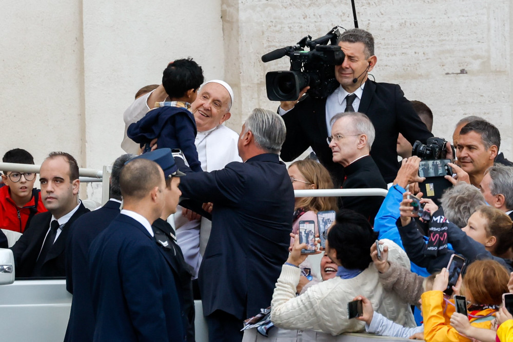 Pope Francis smiles at a child who is held up to him while he is riding in the popemobile. The crowd and a TV cameraman are visible around him.