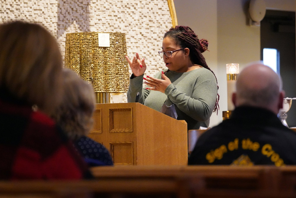 A Black woman wearing glasses signs from behind a wooden lectern. The backs of parishioners in pews watching her are visible.