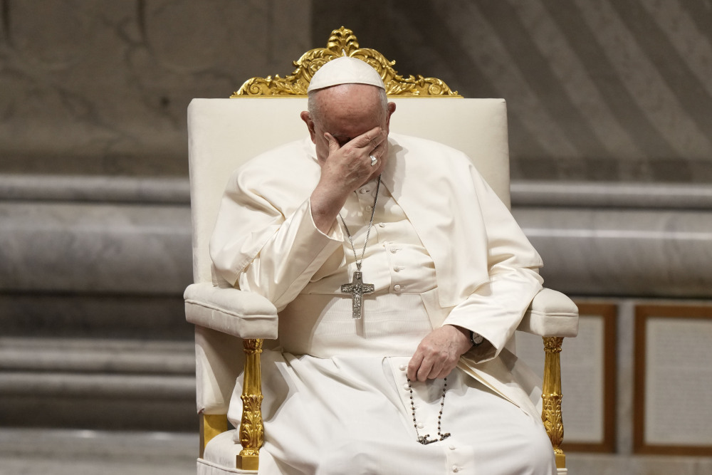 Pope Francis places one hand over his eyes and holds his rosary in the other while seated in a white and gold chair.