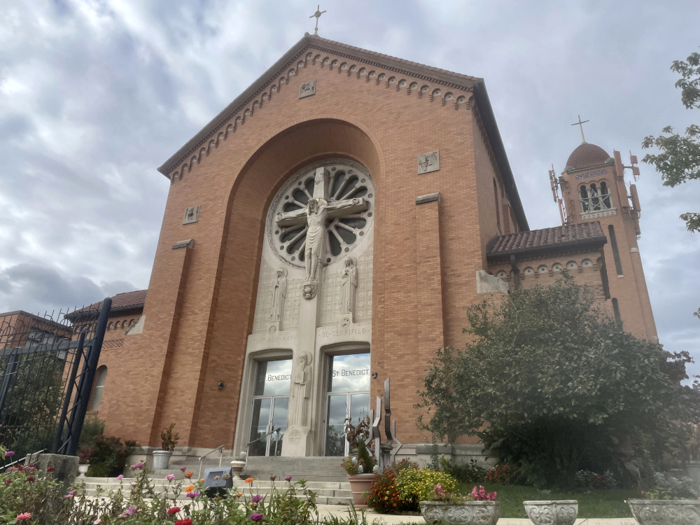 The outside of a high-ceilinged Catholic church with a crucifix on the facade and flowers around the entrance