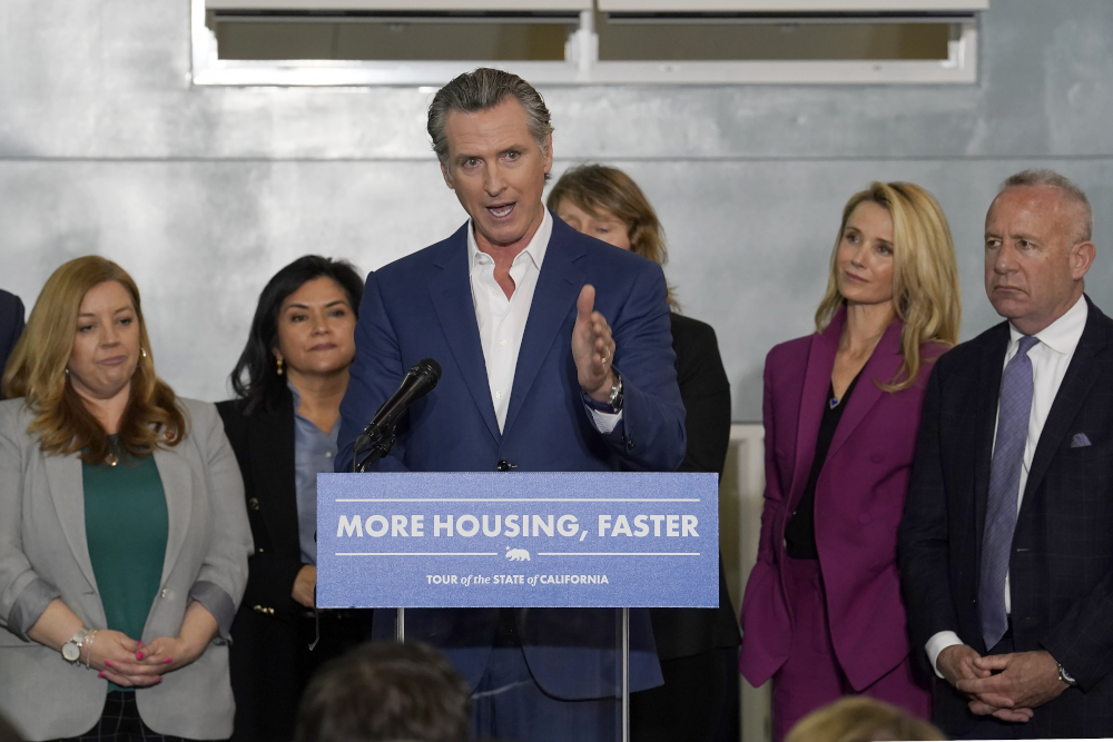 A white man with graying hair wearing suit jacket with no tie speaks from behind a podium that reads "more housing, faster" as others in business professional wear stand behind him