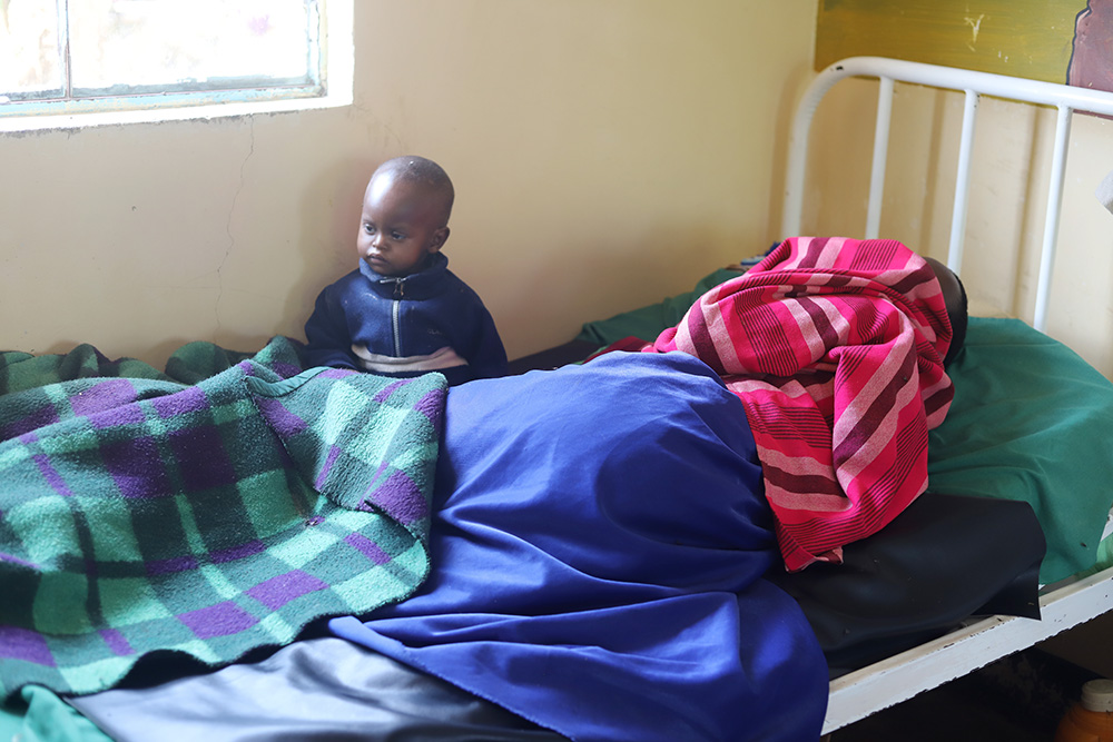 A sick woman sleeps in a hospital bed with her son sitting beside her at Endulen Hospital, located in the Ngorongoro conservation area in northern Tanzania. (GSR photo/Doreen Ajiambo)