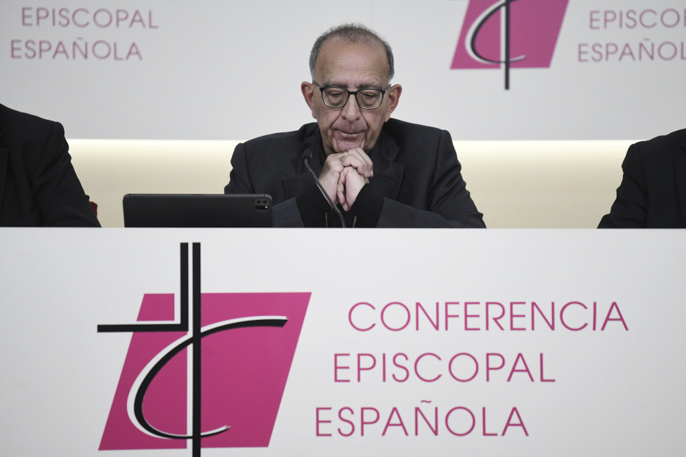 A white man wearing black and glasses closes his eyes and folds his hands as he sits at a table labeled "Conferencia Episcopal Española."