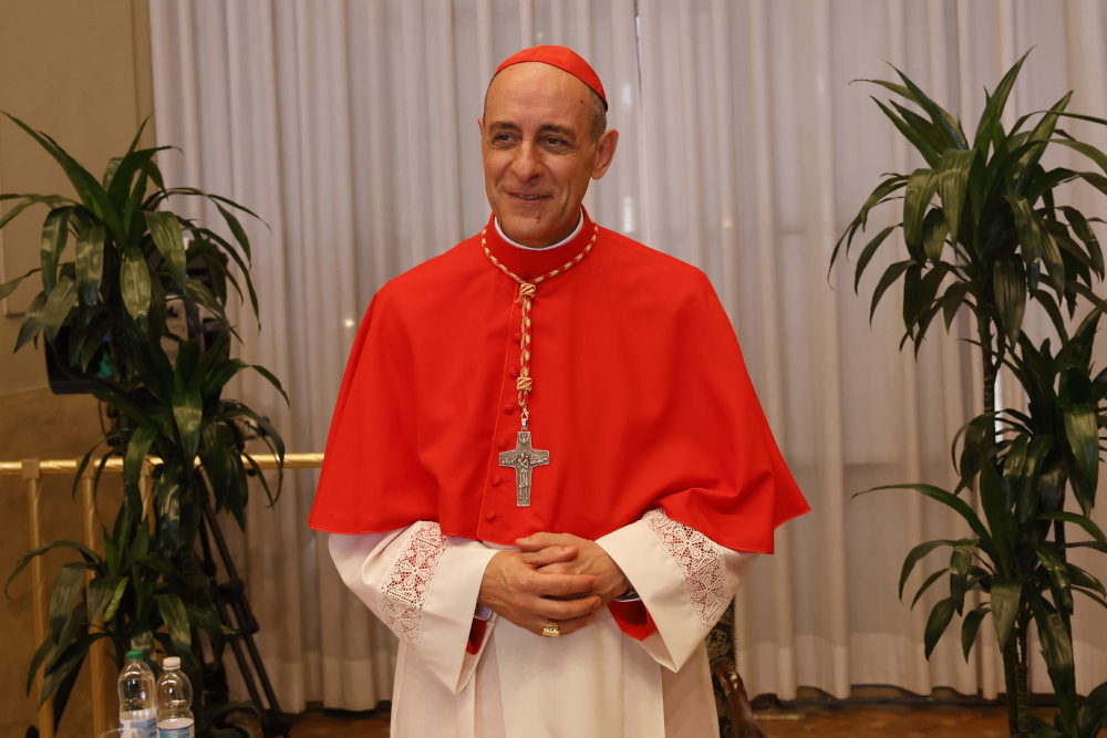 A light-skinned man wears a red cassock and zucchetto and folds his hands in front of him