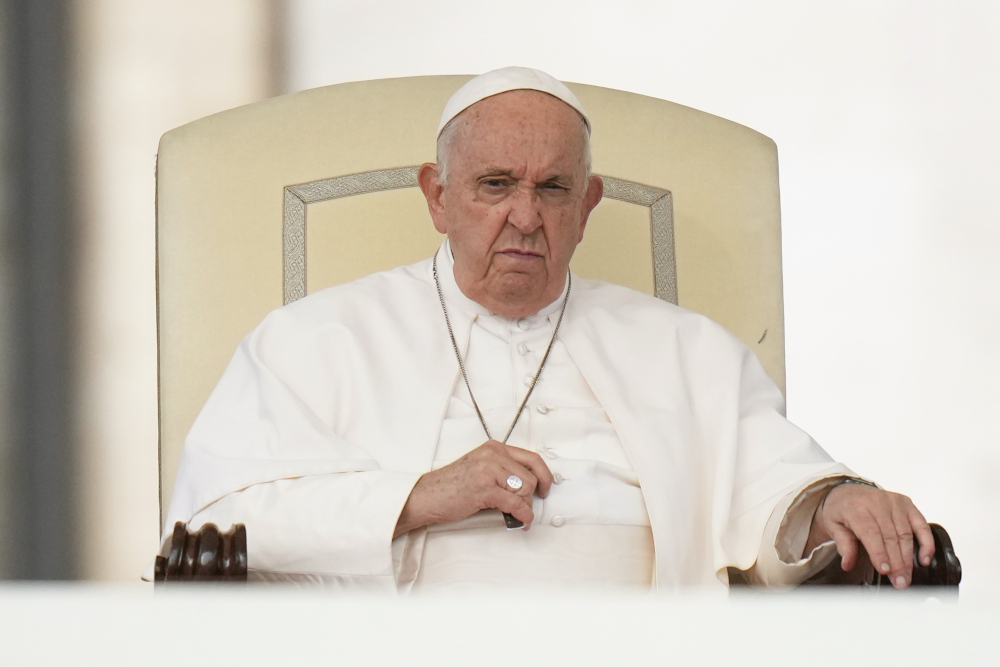 Pope Francis sits in his white chair with a very serious face as his hands close around his pectoral cross
