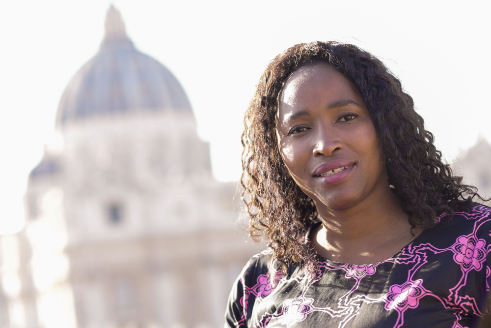 A lighter-skinned Black woman wearing a purple dress looks at the camera with St. Peter's Basilica dome visible in the background