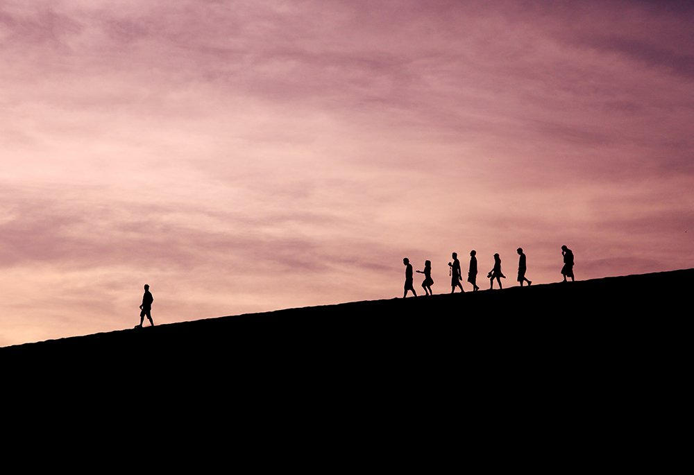 A photo illustration shows a wide shot of a group of people in silhouette, walking along a terrain. One person walking far ahead of the others. (Unsplash/Jehyun Sung)