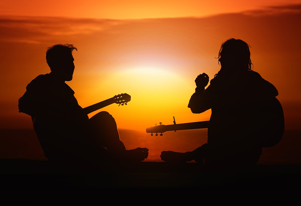 Two musicians play guitar outside, appearing in silhouette against a sunset. (Pixabay/Gerd Altmann)