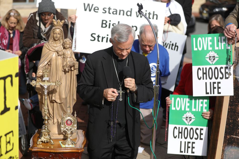 Bishop Strickland holds a rosary. People around him are bowing their heads. A person behind him holds a sign that reads: "We stand with Bishop Strickland."