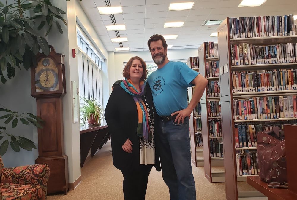 Man and woman stand near bookshelves inside library. 
