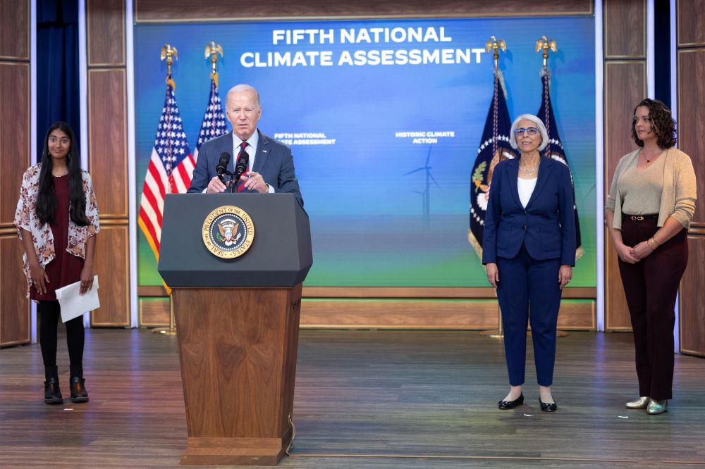 President Joe Biden stands at lectern, speaking, while three women stand nearby. The wall behind him reads, "Fifth National Climate Assessment."