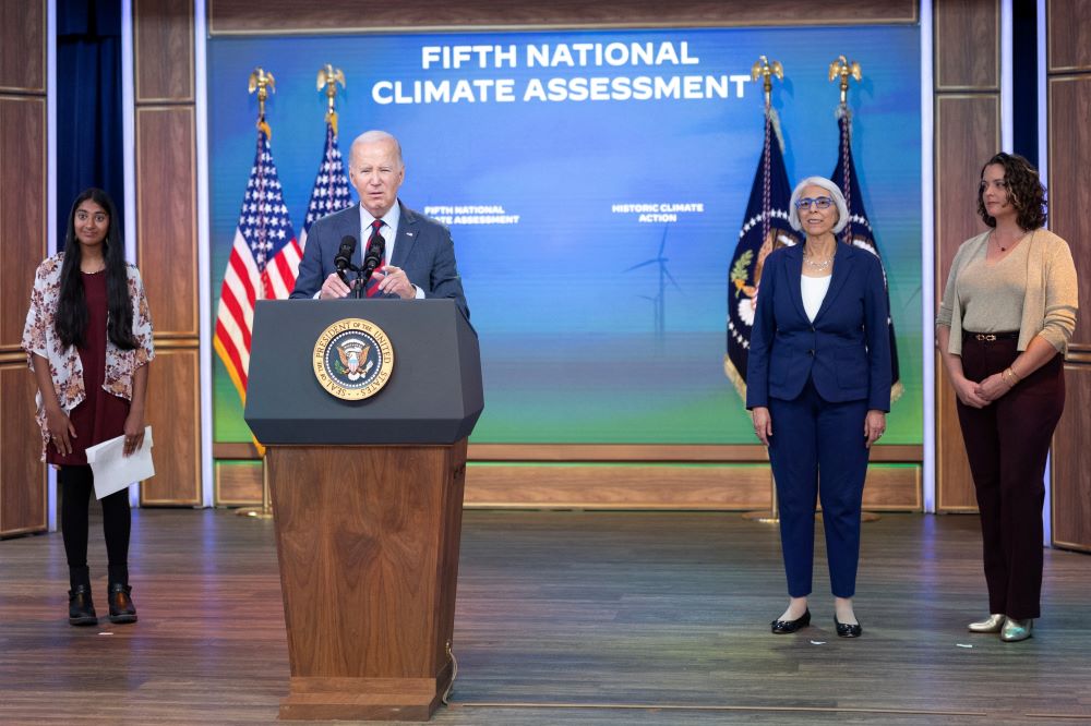 President Joe Biden stands at lectern, speaking, while three women stand nearby. The wall behind him reads, "Fifth National Climate Assessment."