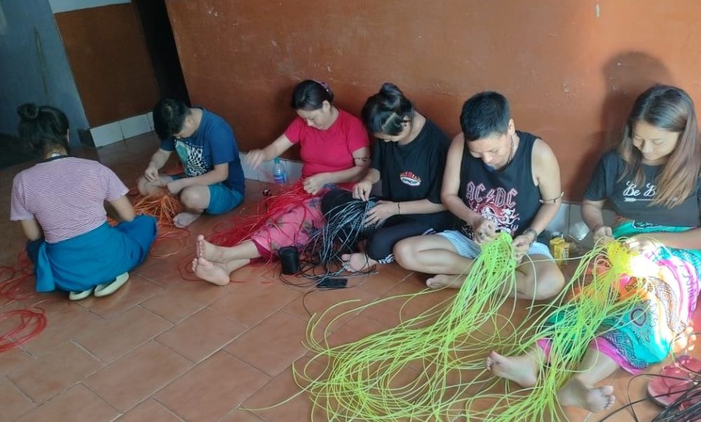 Women sit on the floor, weaving colorful strings.