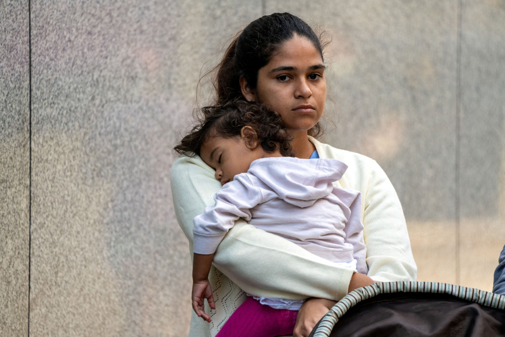A young brown woman stands and holds a sleeping toddler. The woman looks serious.