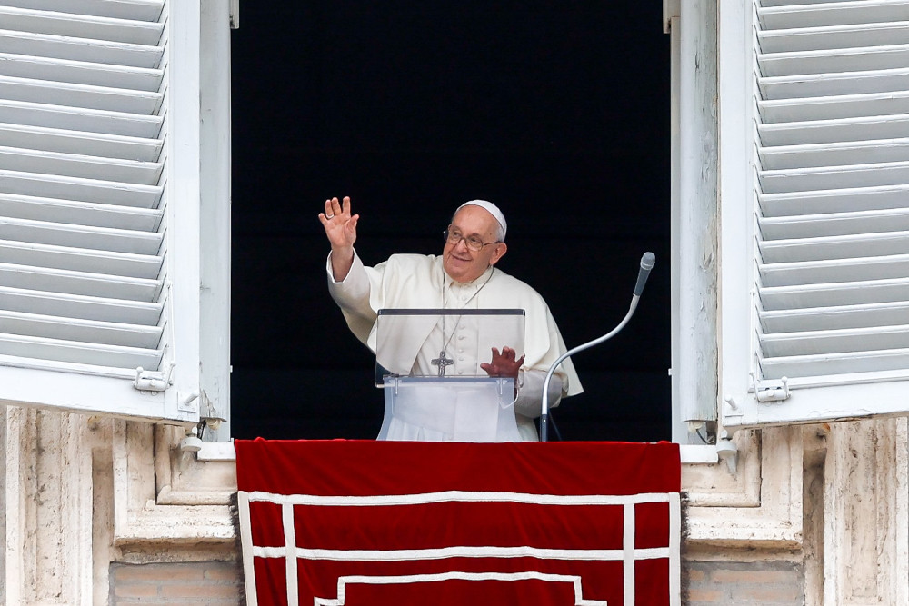 Pope Francis smiles and waves from his apartment window with his other hand on the clear lectern