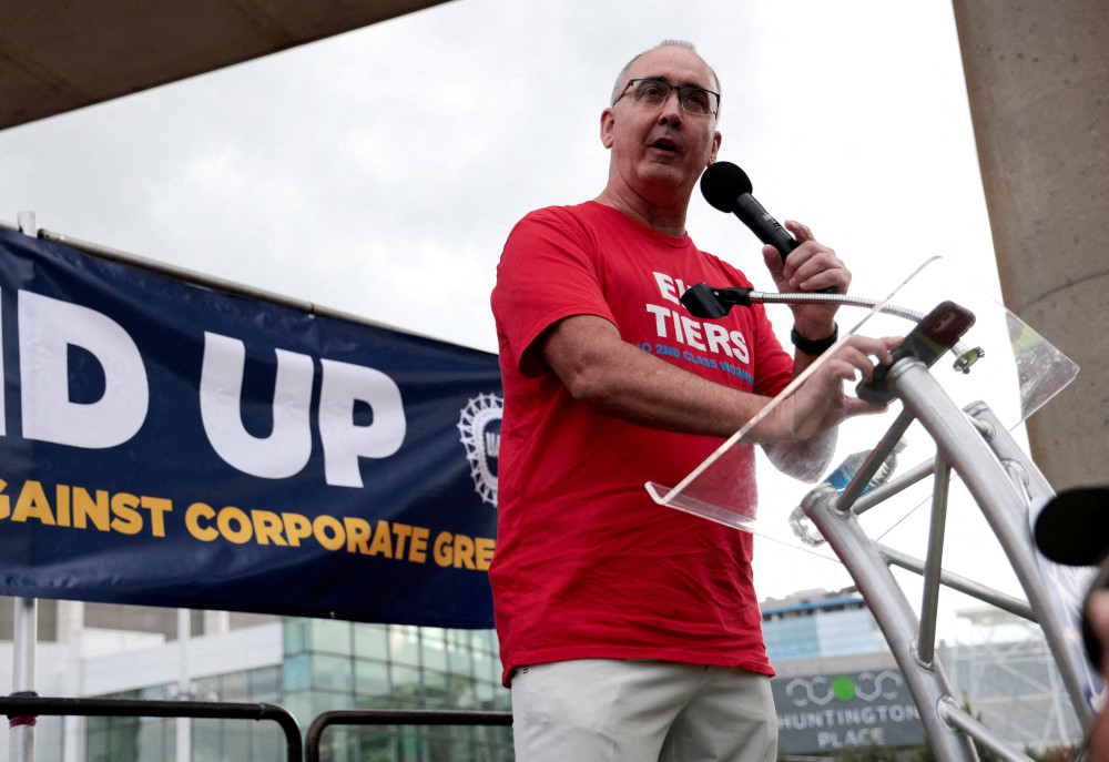 A white man wearing glasses wears a red t-shirt and speaks into a microphone from behind a podium