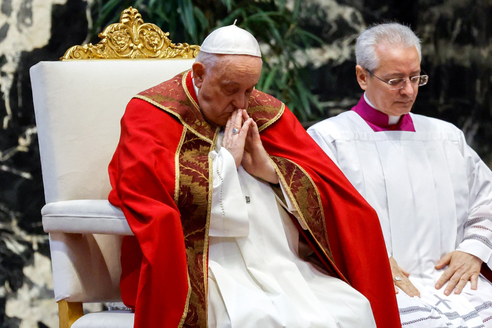 Pope Francis wearing a red cape over his ususal white cassock bows his head and holds his palms together at his chest while sitting in a white and gold chair.
