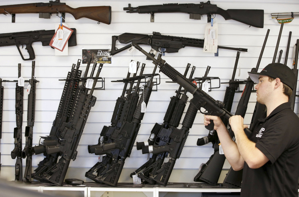 A white man wearing a ball cap holds an AR-15 against his chest. On the wall behind him are many guns, including other AR-15s.