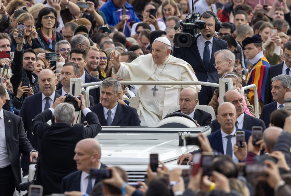 Pope Francis waves at the crowd surrounding his popemobile. A TV camera films him in the popemobile.