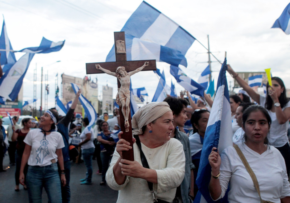 A woman carries a crucifix surrounded by a crowd of other people, many carrying Nicaraguan flags
