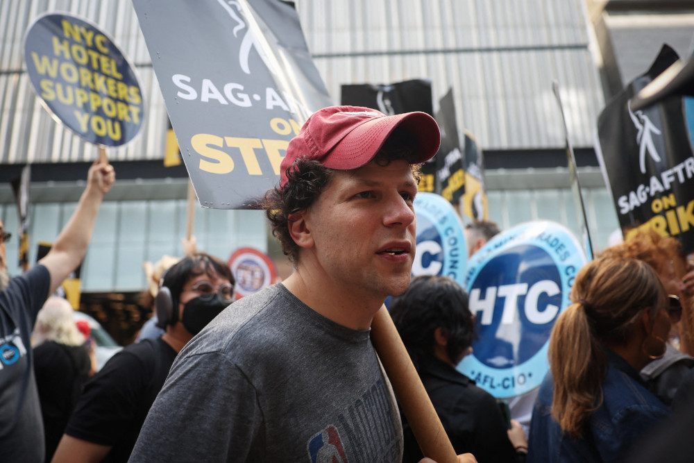 A white man wearing a ball cap has a SAG-AFTRA on strike sign over his shoulder while surrounded by other people with signs outside an office building