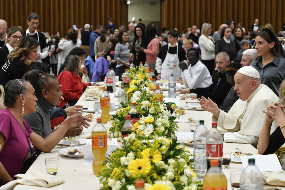 Pope Francis sits at a long table with many other people on both sides. Food, flowers and soft drinks are on the table.