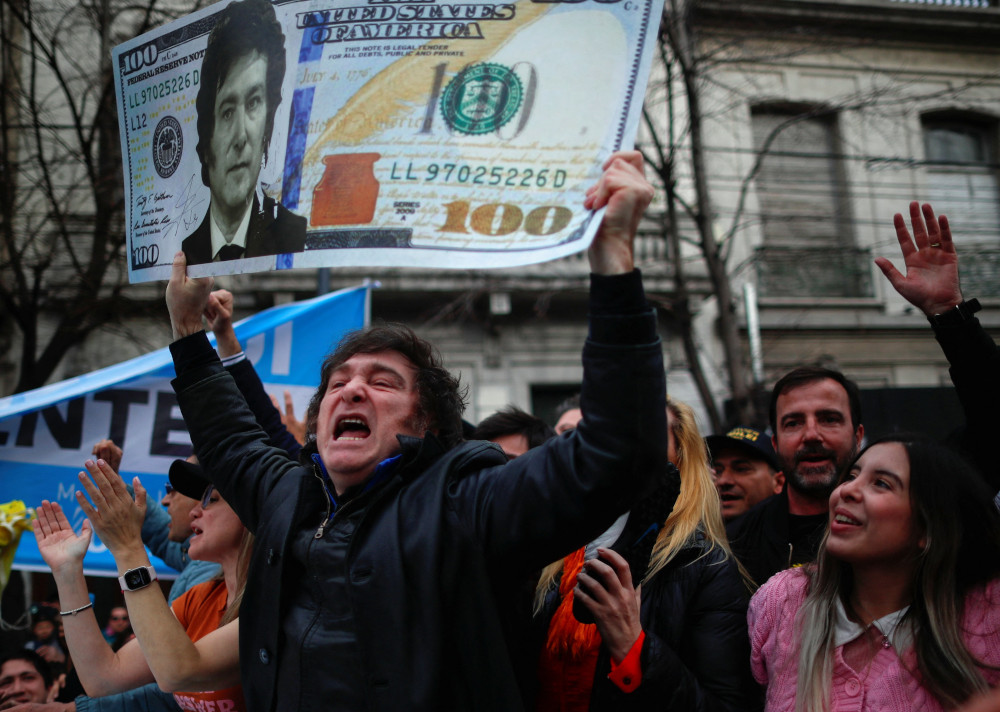 A white man with black hair and sideburns holds a $100 U.S. bill with his face on it, while in a crowd.