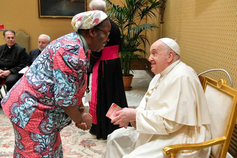 A Black woman wearing a colorful outfit and headscarf leans down to talk to Pope Francis, who is seated