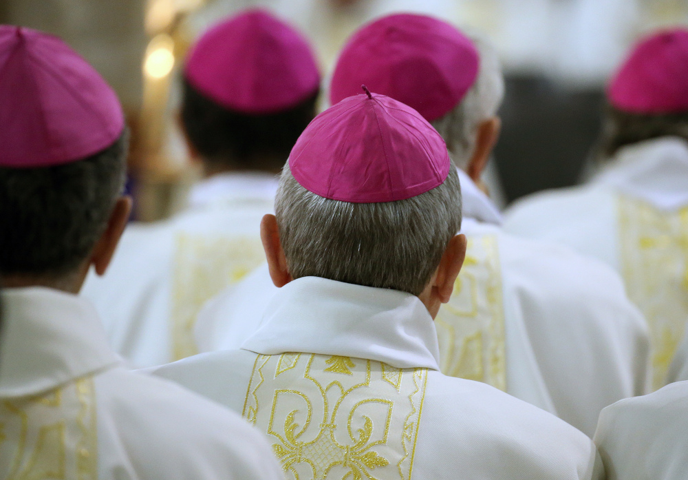 A group of men wearing violet zucchettos and white and gold vestments are visible from behind