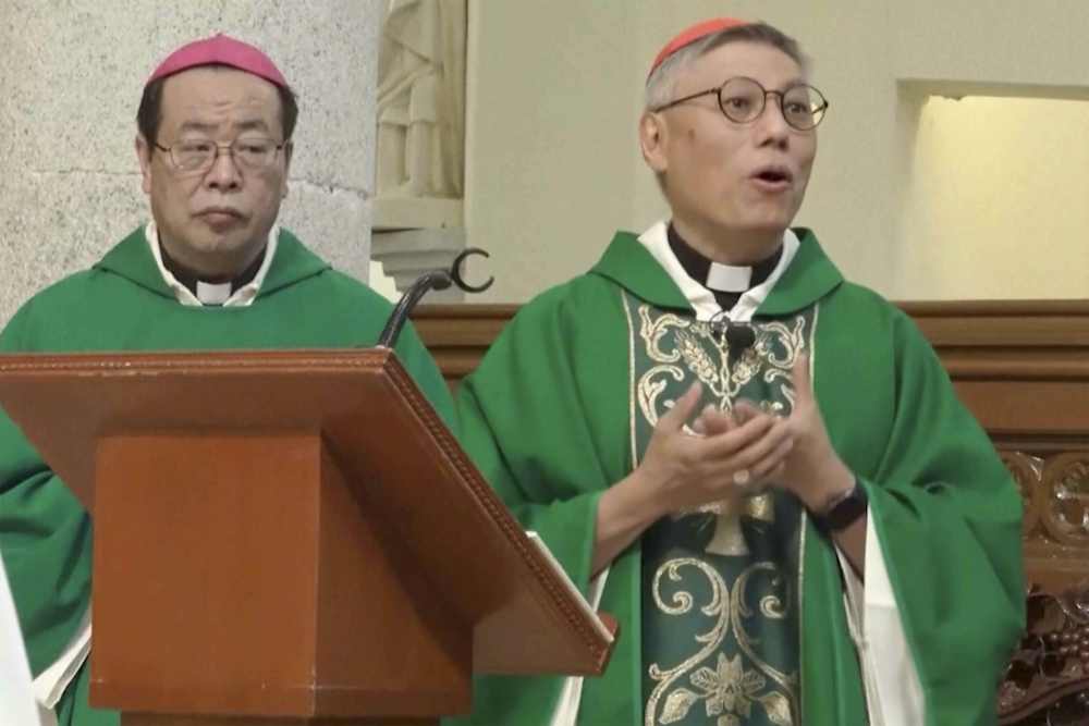 An East Asian man wearing green vestments and a purple zucchetto stands next to another east Asian man wearing green vestments and a red zucchetto. They both stand behind a wooden podium.