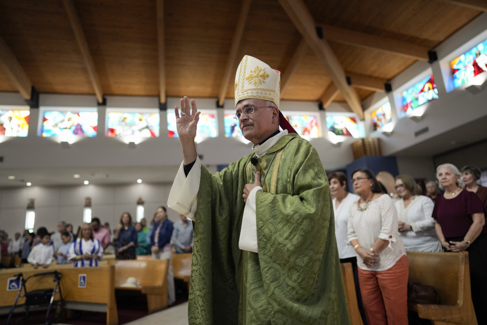A man wearing green vestments and a white mitre raises his right hand while standing before a crowded church