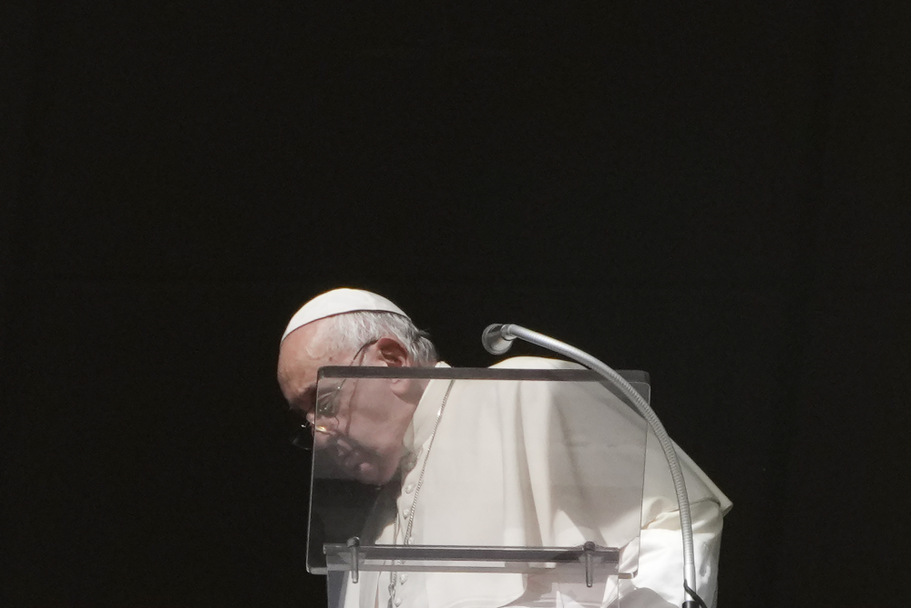 Pope Francis faces the left and looks down from behind a clear lectern with a microphone