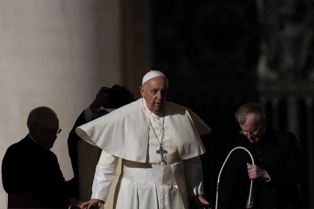 Pope Francis walks with his mozzetta flying out as other men surround him in the shadows