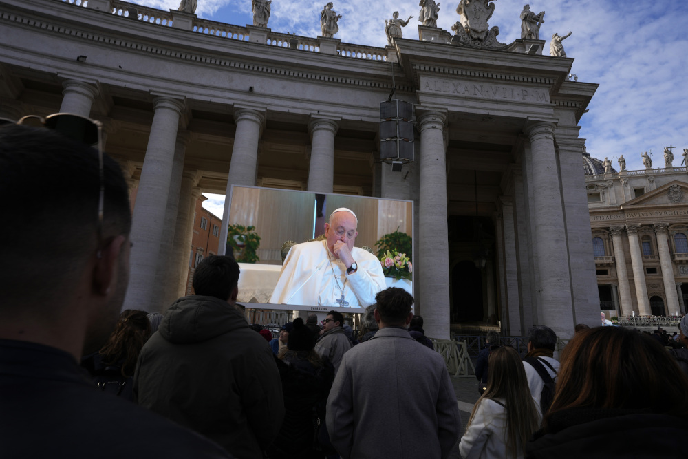 Pope Francis, sitting and holding a fist towards his chin, is projected on a large screen in front of a crowd in St. Peter's Square