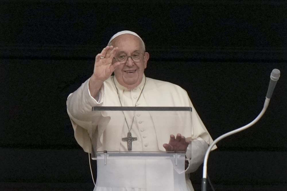 Pope Francis raises his right hand and smiles as he stands behind a clear lectern