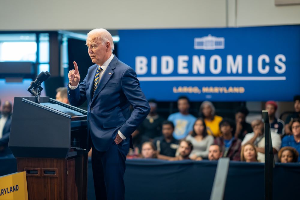 President Biden stands at lectern with people watching him and sign behind him reading "Bidenomics."