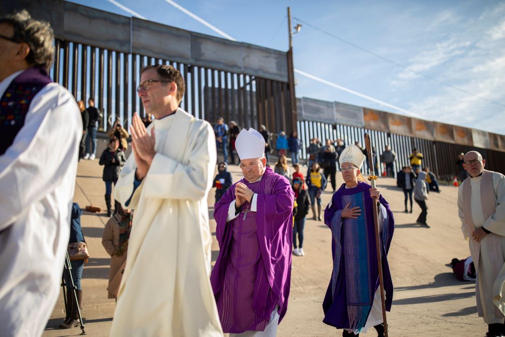 Priests and bishops walk in a line, some praying, with border fence behind them.