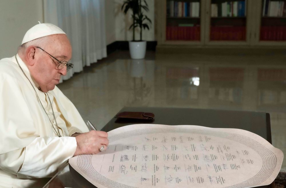 Pope Francis writing on a large, round, white item on a desk.