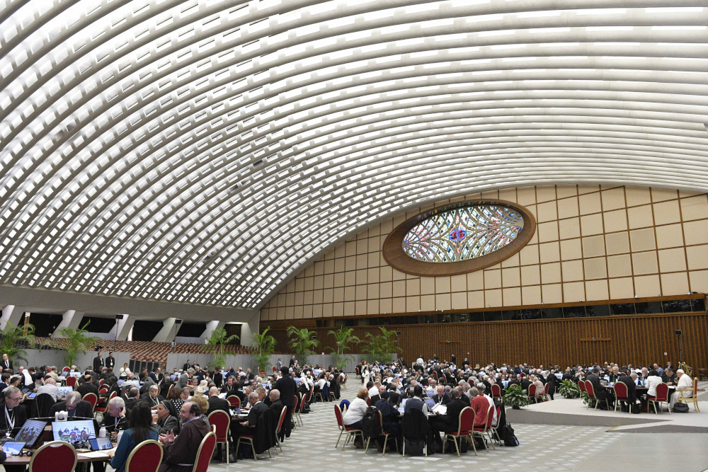 People gather around round tables spread across a large audience hall with an arched roof