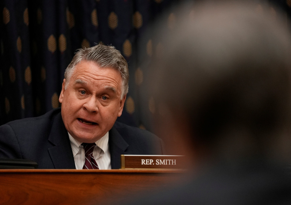 A graying white man wearing a suit and tie speaks next to a Rep. Smith name plate
