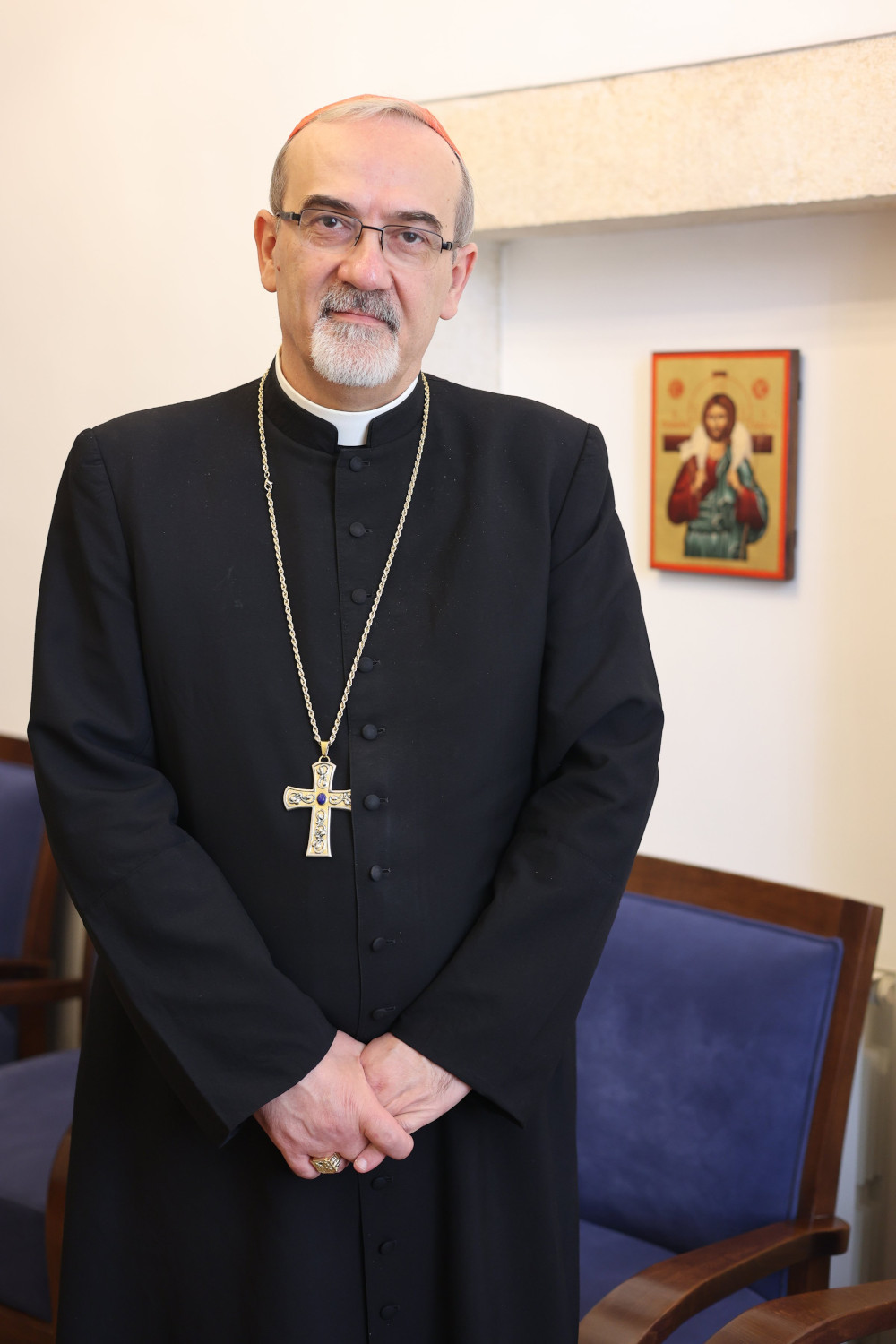 A white man wearing a cassock and a pectoral cross stands and looks at the camera