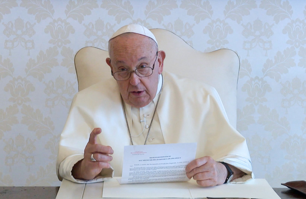 Pope Francis sits in a white chair and points at the camera with a finger as he holds a paper
