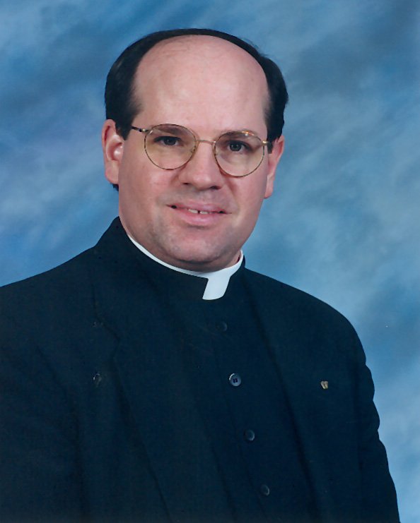 A balding white man with round glasses wears a clerical collar and looks into the camera