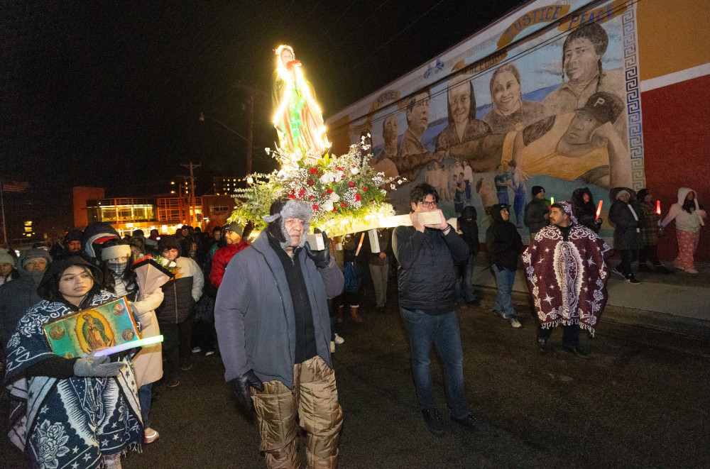 In the center of a crowd outdoors, brown men carry an illuminated Virgen de Guadalupe statue whose base is surrounded by flowers