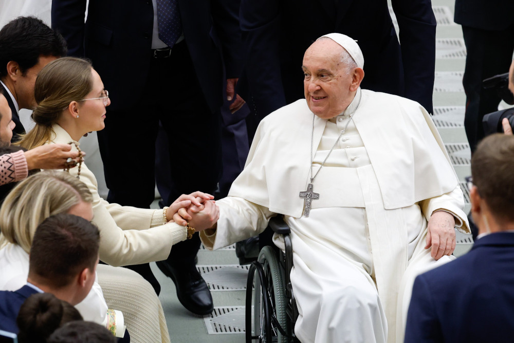 Pope Francis, sitting in a wheelchair, holds the hands of a young woman wearing glasses