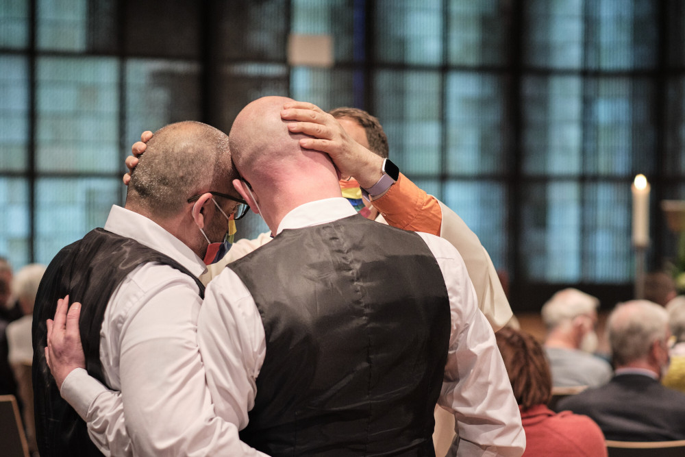 Two men in formal clothes stand close, heads touching, as a priest places his hands on both their heads