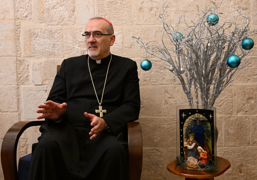 A man wearing a cassock, pectoral cross and red zucchetto sits next to a nativity scene and blue orbs on silvery branches