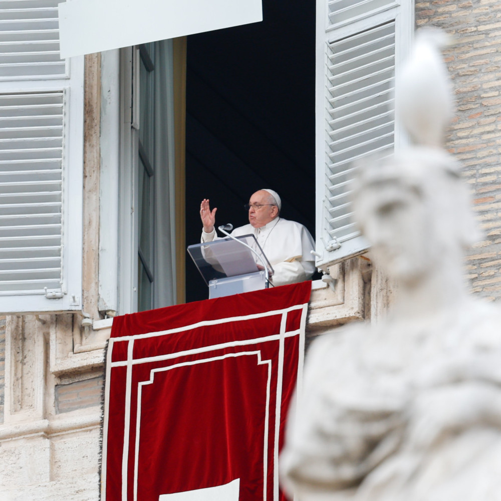 Pope Francis raises his right hand from the window of his apartment. A statue is in the foreground of the photo.