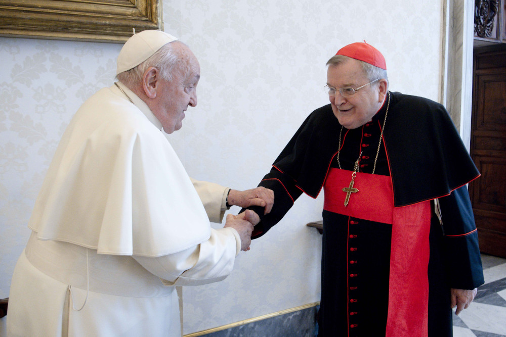 Pope Francis shakes the hand of an older white man wearing a red zucchetto and cardinal's cassock